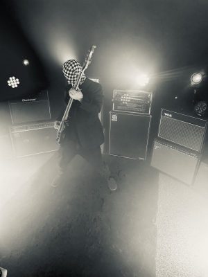 Guitarist in a checkered hood plays a bass on a smoky stage with stacked amplifiers.