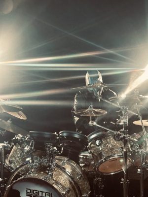 Drummer in a glittery kit under bright stage lights, wearing a hooded jacket behind a sparkling drum set.