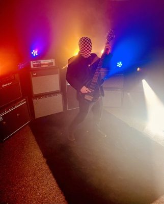 Masked bass player in black, wearing a checkered face mask, performing on a smoky stage with colorful lighting and stacked amplifiers nearby.