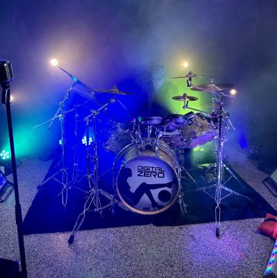 Drum kit on a foggy stage with blue, purple, and green lights; bass drum reads 'DIGITAL ZERO' with a silhouetted figure graphic.