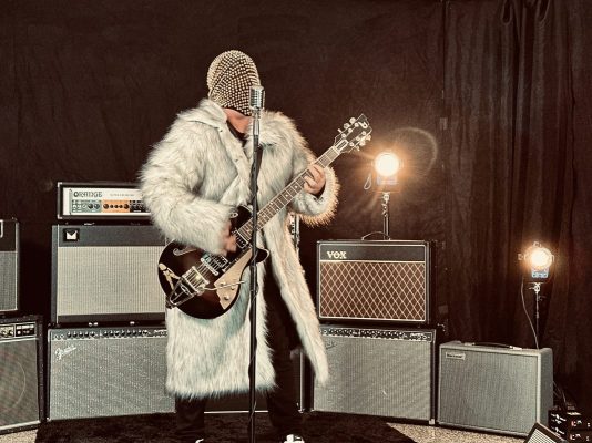 Musician wearing a fluffy white fur coat and beaded mask plays an electric guitar on a stage with multiple amplifiers behind him/her.