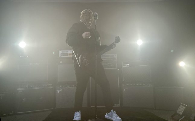 Musician wearing a bead-covered mask on stage, singing into a microphone with a guitar, surrounded by amplifiers and steamy stage fog.
