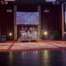Band performing on a dimly lit stage with two guitarists on either side and a drum kit in the center, red curtains behind, and a cityscape mural on the right.