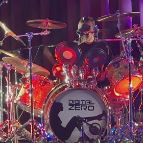 Drummer wearing a plague-doctor style mask behind a shiny drum kit on a purple-lit stage, bass drum labeled DIGITAL ZERO.