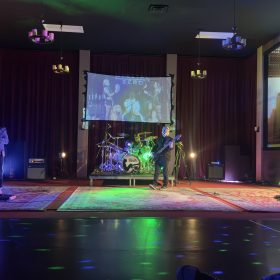 Musicians performing on a lit stage with guitars and drums, a projected screen, and purple curtains in the background.