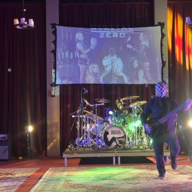 A rock band performs on a lit stage with masked musicians and a large projection screen behind them showing a similar image.