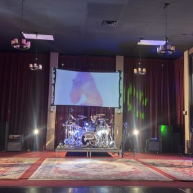 Stage with a live rock band: guitarist left, bassist right, drum kit center, red curtains, and cityscape mural on the wall behind.