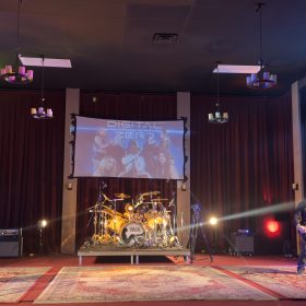 Live band on stage with two guitarists and a drum kit, a projection screen reading 'Digital Zero' behind burgundy curtains.