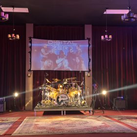 Live rock band on a stage: guitarist left, bassist right, drummer center, red curtains, projection screen reading 'DIGITAL ZERO' above the drum kit with colorful stage lights.