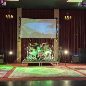 Band performs on stage: two masked guitarists flank a central drum kit; projection screen shows a car interior behind red curtains.