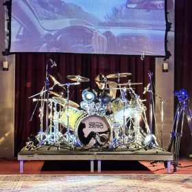 Drum kit on a stage with a large screen projecting an image of a car interior behind it.