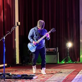 Guitarist wearing a studded mask and blue hoodie, playing a blue electric guitar on a stage with red curtains behind.
