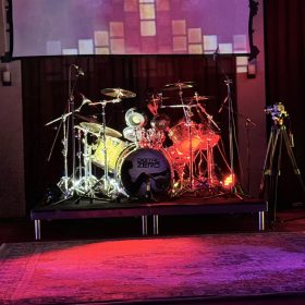 Drum kit on a stage with a large screen showing a colorful grid projection behind it. Purple and red stage lighting accentuates the scene.