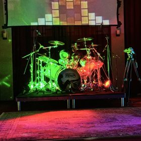 Drum kit on a stage under multicolored lights with a square‑tile projection backdrop behind.