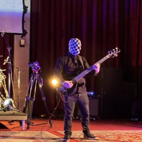 Bassist on stage wearing a black-and-white checkerboard mask, playing a black bass guitar under purple lighting.