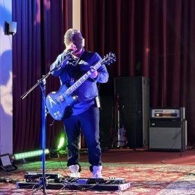 Musician on stage wearing a spiked helmet, singing into a microphone while playing a blue electric guitar with pedal effects at their feet.