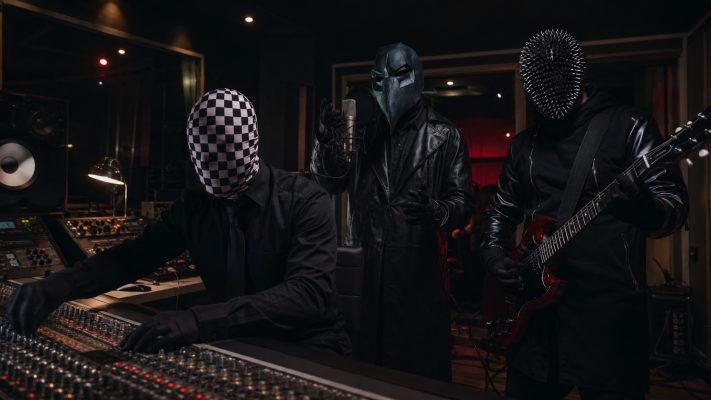 Three masked musicians in a dim recording studio: one at the mixing console, another singing into a microphone, and a guitarist wearing a spiked helmet.